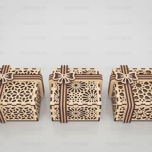 May include: Three square wooden gift boxes featuring intricate laser-cut geometric patterns on the sides and lids. Each box is adorned with a decorative wooden ribbon and bow element in a darker brown tone, contrasting with the light tan wood.