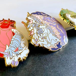 Puede incluir: Tres pines de esmalte con criaturas míticas. El primer pin es un toro rojo y rosa con una cabeza de unicornio blanca. El segundo pin es una nube morada y azul con tres unicornios blancos. El tercer pin es un pin verde y blanco con un unicornio blanco.
