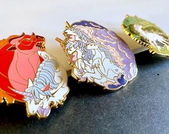 May include: Three enamel pins featuring mythical creatures. The first pin is a red and pink bull with a white unicorn head. The second pin is a purple and blue cloud with three white unicorns. The third pin is a green and white pin with a white unicorn.