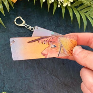 K&ouml;nnte beinhalten: Ein rechteckiger Schl&uuml;sselanh&auml;nger mit einer Illustration eines Profils einer Person mit Fl&uuml;gel. Das Kunstwerk hat eine warme Farbpalette mit Orange-, Gelb- und Rosat&ouml;nen. Der Schl&uuml;sselanh&auml;nger hat einen silberfarbenen Metallring und -verschluss.