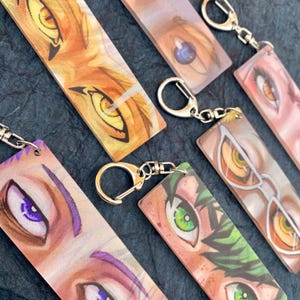 May include: Set of six colorful anime eye keychains. Each keychain features a close-up of a different character's eye with a unique color scheme and style. The keychains are made of acrylic and have a silver metal ring.