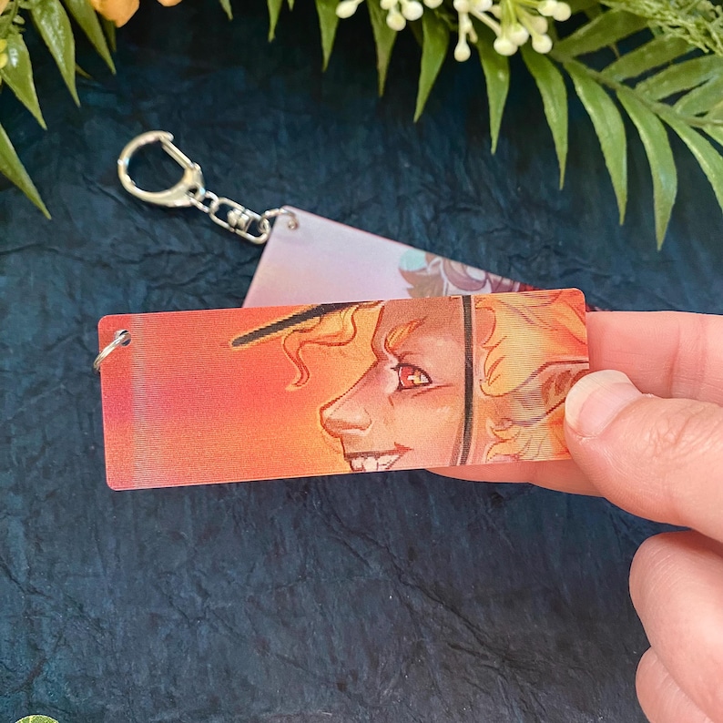 K&ouml;nnte beinhalten: Ein rechteckiger Schl&uuml;sselanh&auml;nger mit einer Charakterillustration vor einem Hintergrund in Sonnenuntergangsfarben. Die Figur hat gelbes Haar und einen schelmischen Gesichtsausdruck. Der Schl&uuml;sselanh&auml;nger hat einen silbernen Schl&uuml;sselring.