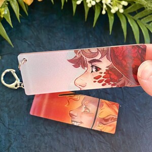 K&ouml;nnte beinhalten: Zwei rechteckige Schl&uuml;sselanh&auml;nger mit Illustrationen von Fantasy-Charakteren. Einer zeigt ein Profil mit roten und braunen Haaren und einem roten Muster. Der andere hat einen Charakter mit gelben Haaren und einem Sonnenuntergangshintergrund. Ein silberner Schl&uuml;sselring ist befestigt.