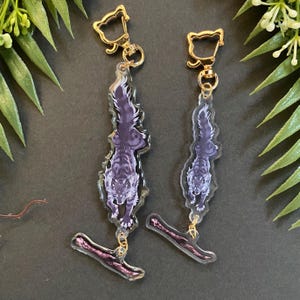 May include: Two keychains featuring a purple wolf design. The wolf is depicted in a stylized, detailed illustration. Each keychain has a gold-colored clasp and a decorative, transparent charm at the bottom. The background is dark.