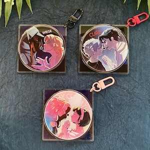 May include: Three clear acrylic keychains with illustrations of couples. Each keychain has a different illustration with the words "WIEGE", "CURE", and "MY CLARITY" on them. The keychains have black, red, and pink clasps.