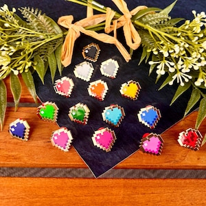 May include: A collection of pixelated heart-shaped enamel pins in various colors, including red, pink, blue, green, and yellow. Each pin has a gold-colored border and a red backing. The pins are arranged on a dark blue surface with decorative greenery.