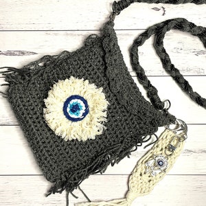 May include: A gray crocheted bag with a white tassel and a white crocheted evil eye charm. The bag has a long black braided strap.