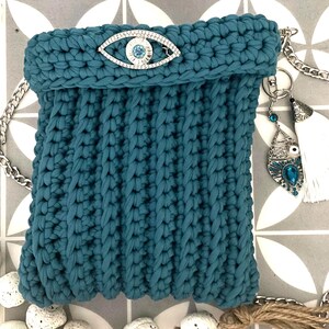 May include: Teal crocheted bag with a silver eye charm and a white tassel. The bag has a chain strap and is decorated with a silver eye charm with blue rhinestones.