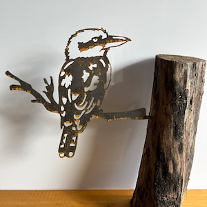 K&ouml;nnte beinhalten: Metallskulptur eines Kookaburra-Vogels, der auf einem Ast sitzt und an einem Teil eines Baumstamms befestigt ist. Der Vogel besteht aus dunklem Metall mit rostfarbenen Akzenten. Der Baumstamm ist dunkelbraun.
