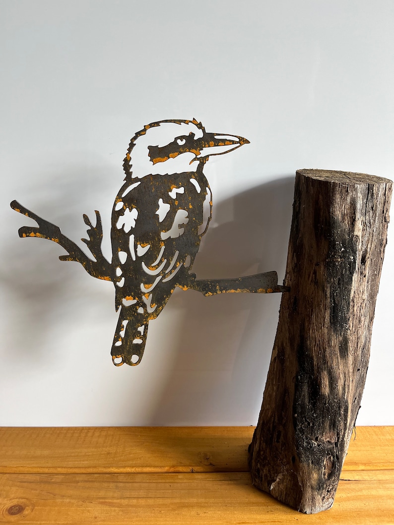 K&ouml;nnte beinhalten: Metallskulptur eines Kookaburra-Vogels, der auf einem Ast sitzt und an einem Teil eines Baumstamms befestigt ist. Der Vogel besteht aus dunklem Metall mit rostfarbenen Akzenten. Der Baumstamm ist dunkelbraun.