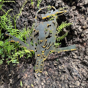 K&ouml;nnte beinhalten: Eine Metallskulptur eines Vogels mit langem Schnabel, der auf einem Ast sitzt. Der Vogel ist aus Metall geschnitten und hat ein verwittertes, rostiges Aussehen und gelbe Akzente. Die Skulptur befindet sich vor einem Hintergrund aus Baumrinde und gr&uuml;nem Laub.
