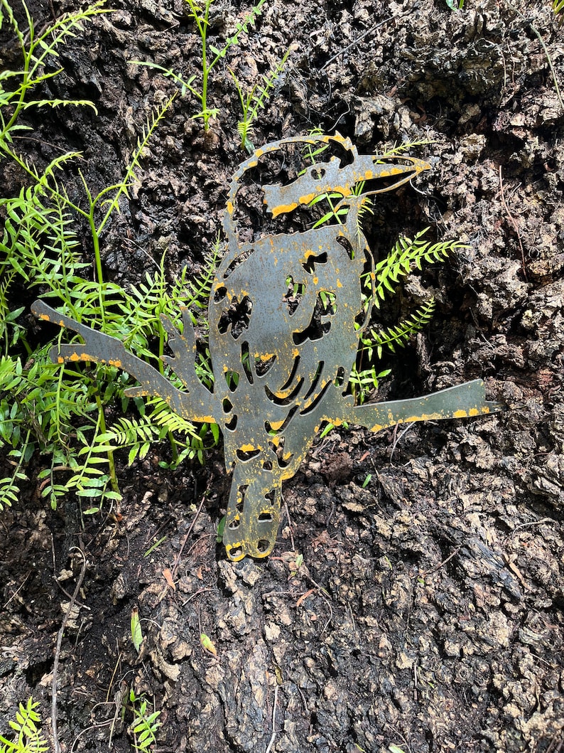 K&ouml;nnte beinhalten: Eine Metallskulptur eines Vogels mit langem Schnabel, der auf einem Ast sitzt. Der Vogel ist aus Metall geschnitten und hat ein verwittertes, rostiges Aussehen und gelbe Akzente. Die Skulptur befindet sich vor einem Hintergrund aus Baumrinde und gr&uuml;nem Laub.