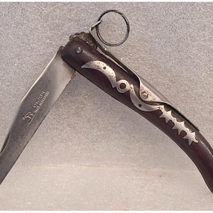 May include: A vintage folding knife with a dark brown wooden handle and a silver blade. The handle features decorative silver accents, including a series of stars. The blade is partially open, revealing the text "Okapi Made in Germany". A small metal ring is attached to the end.