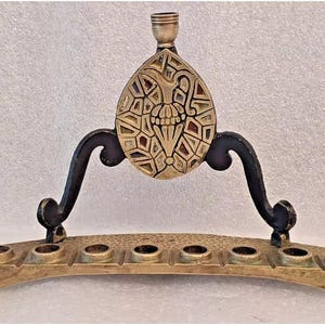 Puede incluir: Una menorá de latón con una base de metal negro y una pieza central decorativa. La menorá tiene siete portavelas y un pico central para aceite.