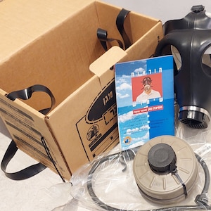 May include: A black gas mask with a filter canister and a set of instructions in Hebrew. The gas mask is in a cardboard box with a black strap.