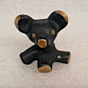 Puede incluir: Pequeña figura de oso koala negro con detalles dorados en las orejas, la nariz y los pies. El koala tiene un diseño sencillo y estilizado y está de pie con los brazos extendidos. La figura está hecha de un material sólido y tiene un acabado mate.