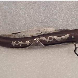 May include: A vintage pocket knife with a dark brown wooden handle. The handle is decorated with silver-colored star and scroll designs. The blade is silver and shows signs of wear. A metal ring is attached to the end of the handle.