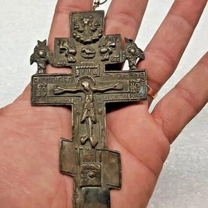 Puede incluir: Una antigua cruz ortodoxa de tono plateado con intrincados grabados e iconografía religiosa. La cruz presenta una representación central de la crucifixión, con inscripciones en un idioma desconocido. La cruz se sostiene en una mano.