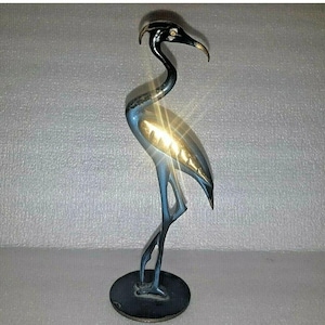 Puede incluir: Escultura decorativa de pájaro de metal. El pájaro tiene un cuello largo y curvado y está sobre una pata. El cuerpo es azul y dorado, con cabeza y pico negros. La escultura está sobre una base redonda.