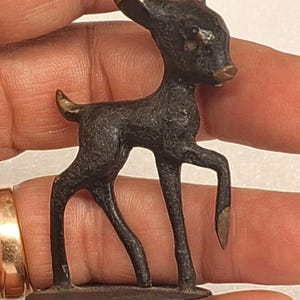 Puede incluir: Pequeña figura de ciervo de color bronce, con una superficie texturizada y detallada. El ciervo tiene un cuerpo esbelto, patas largas y una pequeña base redondeada. Las puntas de las orejas, la cola y la nariz están resaltadas con un acabado dorado. La figura se sostiene en una mano.