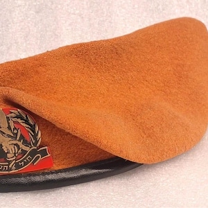 May include: An orange beret with a black leather brim. The beret has a metal emblem with a red and black design and text. The beret is made of a soft, textured material.