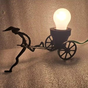 Puede incluir: Una lámpara de metal negro con la silueta de una persona tirando de un carro. La lámpara tiene una bombilla transparente y una base de metal negro.