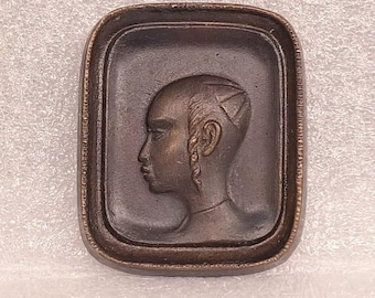 May include: A bronze plaque featuring a raised relief portrait of a person with a braided hairstyle. The plaque is rectangular and has a raised border.