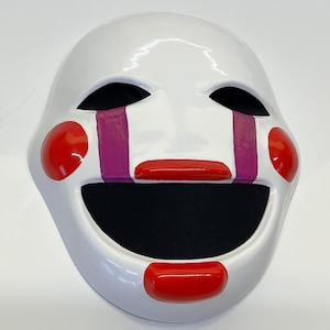 Puede incluir: Una máscara blanca con agujeros negros para los ojos, mejillas rojas y boca roja. La máscara tiene rayas de lágrimas moradas y está diseñada para parecerse a una cara de marioneta o muñeca. La zona de la boca es negra.