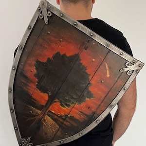Puede incluir: Un escudo de madera con una escena de paisaje pintada que presenta un árbol silueteado contra una puesta de sol. El escudo tiene un marco de metal y detalles decorativos en las esquinas. La obra de arte está en tonos naranja, rojo y marrón.
