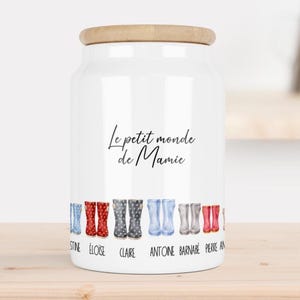 Puede incluir: Un tarro de cerámica blanco con tapa de madera, con el texto "Le petit monde de Mamie" en escritura negra. El tarro está decorado con botas de lluvia de colores y nombres. Un encantador recipiente de cocina.