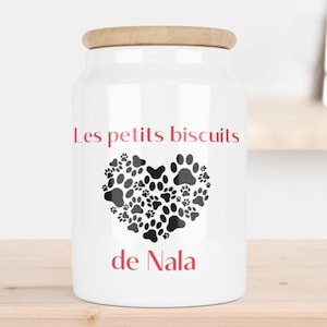 Peut inclure: Pot en céramique blanc avec un couvercle en bois. Le pot porte l'inscription "Les petits biscuits" en rouge, au-dessus d'un motif en forme de cœur composé d'empreintes de pattes noires. Le nom "de Nala" est imprimé en rouge en dessous.