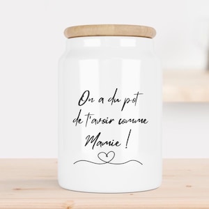 Peut inclure: Un pot de conservation en céramique blanche avec un corps cylindrique et un couvercle en bois clair. L'avant du pot présente un texte noir en écriture cursive "On a du pot de t'avoir comme Mamie !" au-dessus d'un petit cœur dessiné.