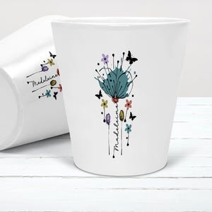 Puede incluir: Dos macetas de cerámica blanca. Una maceta está en posición vertical, la otra está inclinada sobre su lado. Cada maceta presenta un diseño floral turquesa con mariposas y el nombre "Madeleine" en escritura negra. Las macetas tienen un acabado brillante.