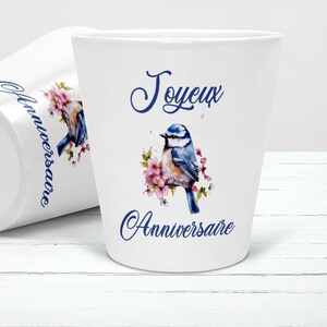 Peut inclure: Pot de fleurs en céramique blanche avec une illustration à l'aquarelle d'un oiseau bleu perché sur des fleurs roses. Le pot porte l'inscription "Joyeux Anniversaire" en écriture bleue. Un pot est debout, l'autre est couché.