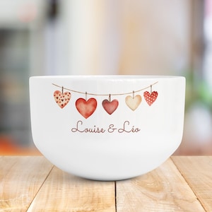 Peut inclure: Un bol en céramique blanc orné d'un motif horizontal représentant une cordelette avec cinq cœurs suspendus à l'aquarelle dans des tons rouge, rose et crème. Sous les cœurs, le texte "Louise & Léo" est écrit en écriture cursive brune.