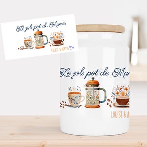 Peut inclure: Un pot en céramique blanc avec un couvercle en bois, orné d'un motif aquarelle de tasses à café, d'une cafetière à piston et d'éléments floraux. Le texte "Le joli pot de Mamie" et "LOUISE & AGATHE" est écrit sur le pot.