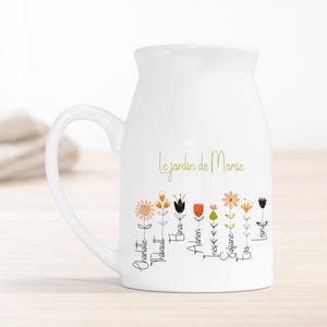 Peut inclure: Mug en céramique blanche avec une anse incurvée. Le mug présente le texte "Le jardin de Mamie" en vert, avec un motif floral orange, noir et rouge. Des noms sont écrits sous les fleurs.