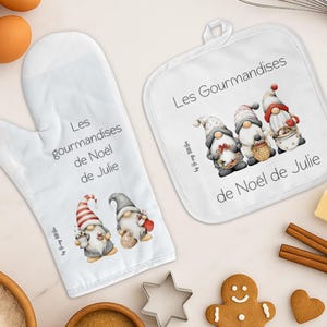 Peut inclure: Un ensemble de manique et de gant de cuisine blancs avec des illustrations de gnomes et le texte "Les gourmandises de Noël de Julie". L'ensemble comprend un biscuit en pain d'épices, des emporte-pièces en forme d'étoile et de cœur, et d'autres fournitures de pâtisserie.