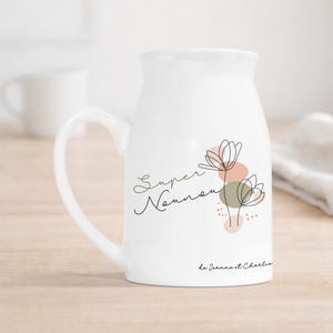 Puede incluir: Una jarra de cerámica blanca con un diseño floral de líneas negras, formas abstractas en color melocotón y verde salvia, y el texto "Super Nounou" junto a "de Jeanne et Charlie" impreso en letra cursiva en la parte frontal.