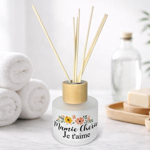 May include: A frosted glass essential oil diffuser with a wooden cap and reeds. The diffuser has a floral design and the text "Mamie Cherie Je t'aime". White towels and a bottle are in the background.