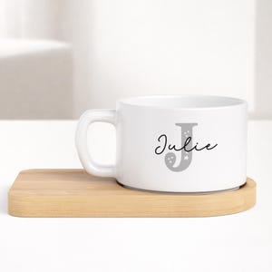 Può includere: Tazza in ceramica bianca con manico, con una lettera "J" grigia e il nome "Julie" in corsivo. La tazza è appoggiata su un sottobicchiere in legno chiaro. Il design è semplice ed elegante, adatto all'uso quotidiano.