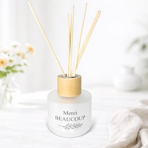 May include: A frosted glass aromatherapy diffuser with a wooden cap and reeds. The diffuser has the words "Merci Beaucoup" and a leaf design printed on the front. The background is a soft white with a vase of flowers.
