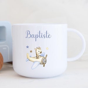 Peut inclure: Une tasse en céramique blanche avec une anse bleue. La tasse est décorée d'une illustration bleue et blanche d'une girafe pilotant un avion avec des étoiles. Le texte "Baptiste" est imprimé en bleu sur la tasse.