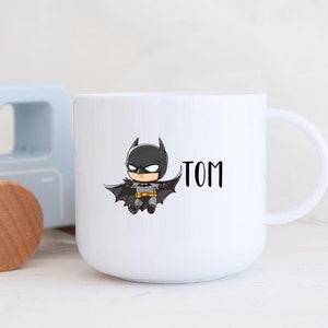 Op de afbeelding: Witte keramische mok met een cartoon afbeelding van Batman en de naam "TOM" erop gedrukt.