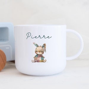 May include: White ceramic mug featuring a bunny illustration and the name "Pierre". A light blue wooden toy truck is in the background. The bunny wears a patchwork outfit. The mug has a handle.