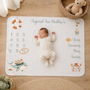 May include: A white milestone blanket featuring nautical illustrations including a lifebuoy, a whale with a village on its back, a crab, and a message in French reading Aujourd'hui Mathis a. The blanket includes numbers 1 through 12 and time units.