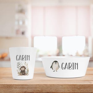 Op de afbeelding: Witte keramische beker en kom set. De beker heeft de naam "GABIN" in grijze letters, samen met een apenillustratie. De kom heeft ook de naam "GABIN" en een pinguïnillustratie. De set staat op een houten oppervlak.