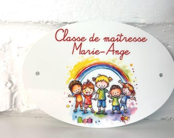 May include: Oval white sign with the text "Classe de maîtresse Marie-Ange" in red script. The sign features a colorful illustration of children under a rainbow. Two small holes are visible for mounting.