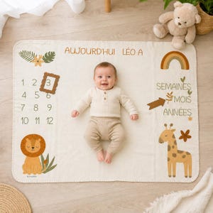 Puede incluir: Una manta de hitos para bebés de color crema con el texto AUJOURD'HUI LÉO A, los números del 1 al 12 y las palabras SEMAINES, MOIS, ANNÉES. La manta incluye ilustraciones de un león, una jirafa, un arcoíris y un marco marrón de marcador.