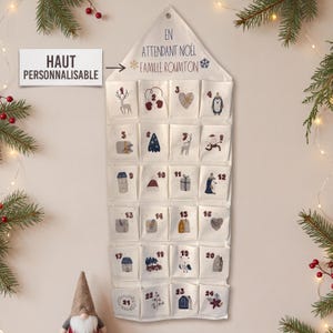 Peut inclure: Un calendrier de l'Avent en tissu crème avec vingt-quatre poches numérotées. La partie supérieure porte l'inscription EN ATTENDANT NOËL FAMILLE ROUMTON. Chaque poche présente une illustration de Noël unique, incluant des rennes, des pingouins, des maisons et des sapins dans des tons doux.
